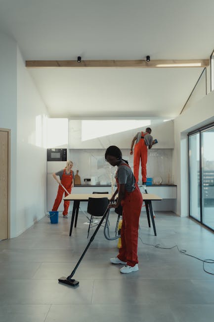 A modern, spacious kitchen with light-colored tiled flooring and white walls illuminated by natural light from large floor-to-ceiling windows on the right. The ceiling features a wooden beam with black spotlights. Three professional cleaners from Cleaners Mayfair, dressed in grey shirts and red uniforms, are engaged in deep cleaning activities: one vacuuming the tiled floor with a cordless vacuum in the foreground, another wiping the work surface with a cloth near the back, and a third cleaning the upper wall area with a step stool. The kitchen has minimalist black chairs around a rectangular table, and the surfaces appear clean and well-maintained, reflecting effective hygiene and surface cleaning practices.