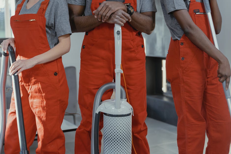 Close-up view of three professional cleaning workers dressed in grey shirts and bright orange overalls, standing indoors with a modern, clean ambiance. The workers are holding vacuum cleaners with silver metal handles and grey hoses, actively engaged in surface cleaning. The background shows a clean, tiled floor, a metal chair, and a black countertop, all well-lit by natural light that highlights the shiny, dust-free surfaces. The image emphasizes thorough hygiene and proper equipment usage, illustrating deep cleaning practices, with Cleaners Mayfair specializing in domestic cleaning and surface sanitisation. The scene reflects a focus on maintaining pristine environments through professional cleaning processes.