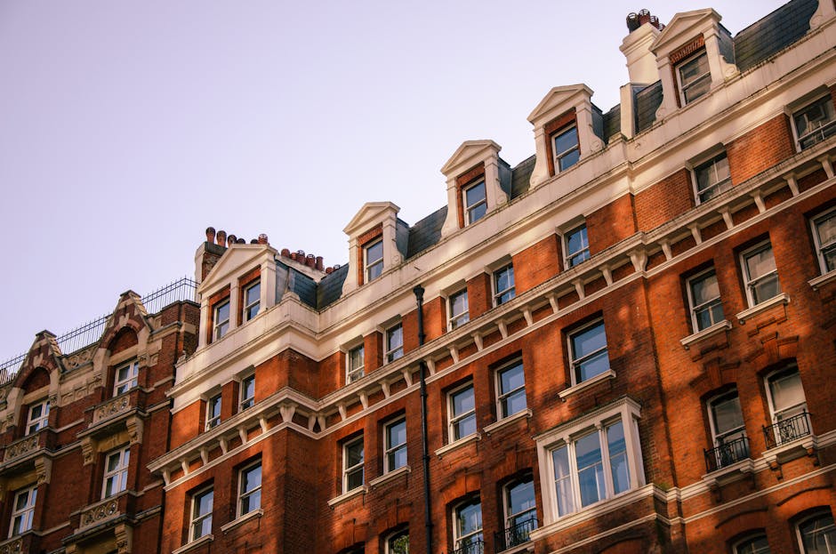 Close-up of the upper floors of a historic red brick residential building in Mayfair, W1K, with detailed white cornices, dormer windows, and black metal railings on the small balconies, illuminated by natural daylight against a clear sky. The building features a classic architectural style with decorative stonework and multiple chimney stacks visible on the roof, indicating an older yet well-maintained structure. This image aligns with the context of domestic and surface cleaning for heritage apartments, exemplifying the importance of deep cleaning and maintenance of exterior surfaces in luxury flats, as provided by Cleaners Mayfair.