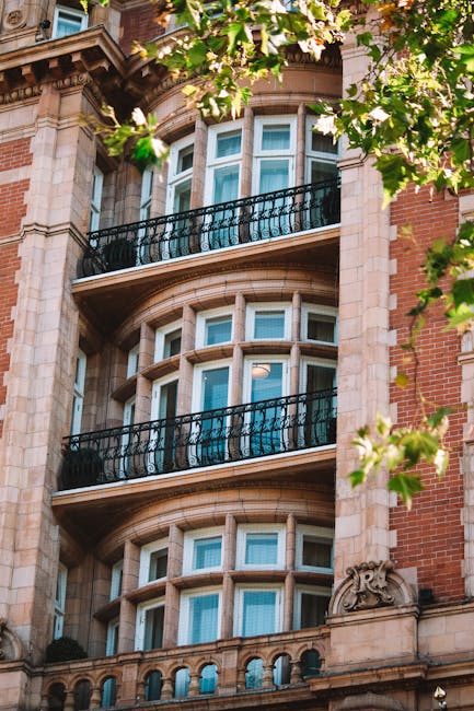 Exterior view of a historic residential building on Park Lane, Mayfair W1K, featuring classic architectural details with large, multi-pane bay windows and ornate stonework. The building’s façade combines red brick with cream-colored stone accents, and black wrought iron balcony railings are visible on the upper floors. Surrounding green foliage partially frames the view, with sunlight illuminating the structure, highlighting its clean, well-maintained surface. This image represents the importance of surface cleaning and maintenance for preserving the aesthetic appeal of luxury flats, aligning with the services offered by Cleaners Mayfair for deep cleaning and sanitisation of residential properties in this prestigious area.