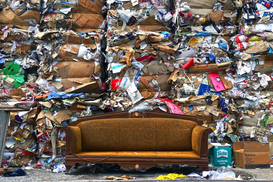 An outdoor scene featuring an old wooden sofa with patterned upholstery placed against a backdrop of a large pile of assorted bulky waste and discarded packaging materials, including cardboard boxes, paper, and plastic wraps. The sofa shows signs of wear, with some minor damage to the legs and fabric. The pile of waste appears messy and unorganized, consisting of various materials such as flattened cardboard, crumpled paper, and plastic packaging, suggesting improper disposal or accumulation in need of waste removal services. The area is well-lit, with natural daylight clearly illuminating the scene, highlighting the contrast between the vintage furniture and the cluttered debris. This image reflects the importance of proper waste management and domestic cleaning, as offered by Cleaners Mayfair, especially regarding bulky waste and mattress disposal.