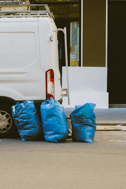A white commercial van parked on a street in front of a building with large glass windows. The van's rear door is partially visible, and three blue garbage bags are placed on the pavement next to the vehicle, indicating waste collection or disposal activity. The street surface appears clean with a smooth asphalt finish. The building's exterior features a beige wall and a glass door, suggesting a commercial or retail space. Natural daylight illuminates the scene, highlighting the cleanliness and orderliness typical of professional waste disposal services. This setting aligns with surface and waste management procedures, as well as the sanitation standards promoted by Cleaners Mayfair for residential and commercial cleaning needs, including bulky waste and mattress disposal. The image focuses on waste collection at a clean, urban environment, emphasizing professional hygiene and disposal processes.