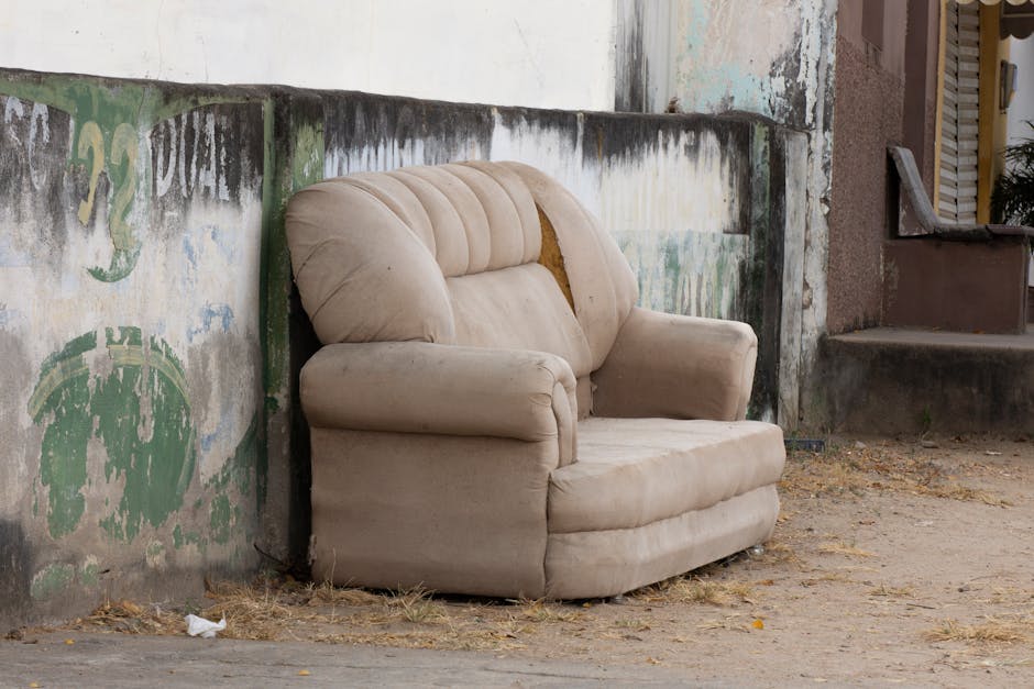 An outdoor scene featuring an old wooden sofa with patterned upholstery placed against a backdrop of a large pile of assorted bulky waste and discarded packaging materials, including cardboard boxes, paper, and plastic wraps. The sofa shows signs of wear, with some minor damage to the legs and fabric. The pile of waste appears messy and unorganized, consisting of various materials such as flattened cardboard, crumpled paper, and plastic packaging, suggesting improper disposal or accumulation in need of waste removal services. The area is well-lit, with natural daylight clearly illuminating the scene, highlighting the contrast between the vintage furniture and the cluttered debris. This image reflects the importance of proper waste management and domestic cleaning, as offered by Cleaners Mayfair, especially regarding bulky waste and mattress disposal.