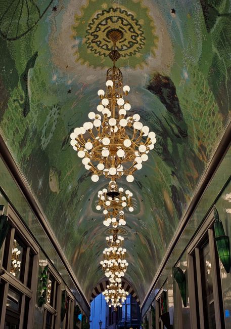 Interior corridor with an ornate, arched ceiling painted with vibrant, abstract artwork, illuminated by a series of large, gold-colored chandeliers featuring multiple spherical white glass shades. The
