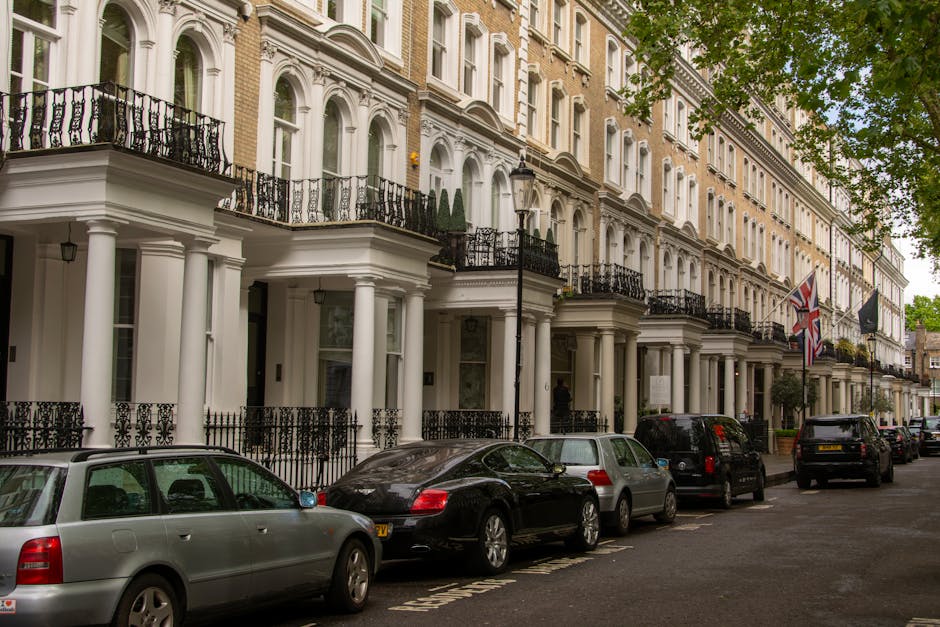 Street scene in Mayfair during early evening with a red classic sports car parked on the curb, surrounded by pedestrians walking along the pavement. The buildings feature a mix of modern and historic 