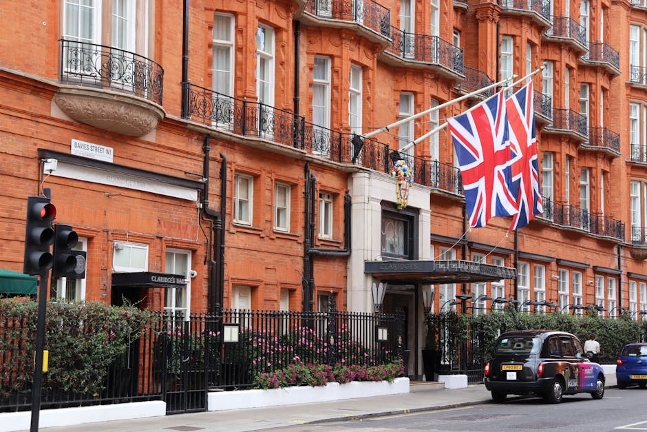Exterior view of the historic Savile Row hotel in Mayfair, featuring a red brick facade with multiple windows framed by white trims, situated behind a black wrought iron fence with decorative elements. British flags are prominently displayed above the entrance, which is marked by a black awning with the hotel's name, 'Claridge's.' The sidewalk in front shows a traffic light and a black taxi parked nearby, with a small person walking on the street. The scene is well-lit with natural daylight, highlighting the building's classic architecture and well-maintained appearance, reflecting the luxurious and elegant atmosphere of the Mayfair district. The image exemplifies the exterior environment where cleaning and maintenance of historic buildings might be relevant, emphasizing pristine upkeep as part of professional cleaning services offered by Cleaners Mayfair.