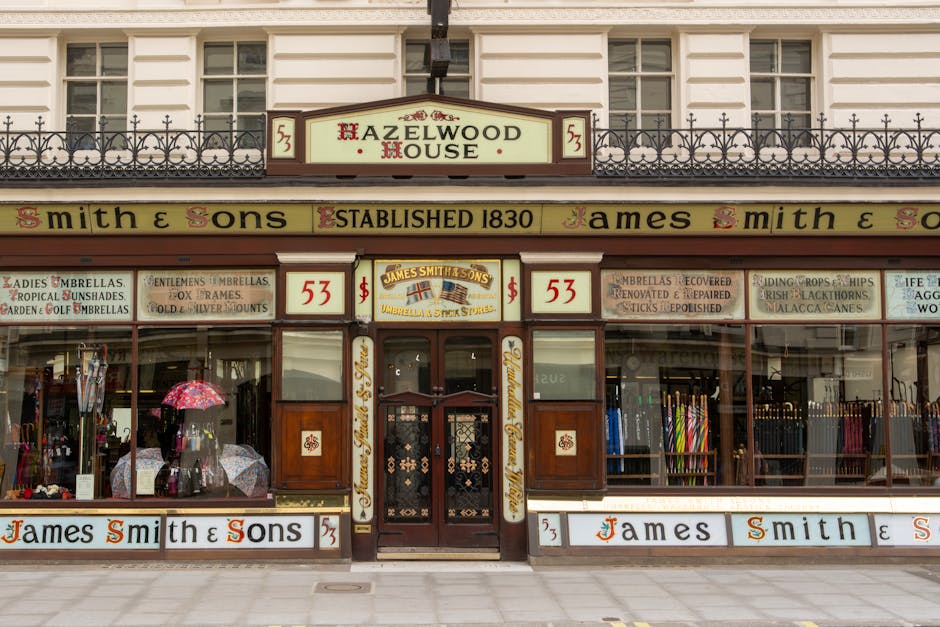 The image depicts the facade of a historic building on Savile Row, featuring a combination of red brickwork and white decorative stone accents. The centered entrance has a black wooden door with glass panels and is framed by an elegant white arch with ornate scrollwork detailing. Above the door, a large window with black framing is visible, accompanied by a small balcony with black wrought iron railings adorned with greenery. The exterior surface appears clean and well-maintained, with no visible dust or dirt. The building's surrounding sidewalk is edged by a black metal fence, and a potted topiary plant is positioned near the doorway, contributing to the building’s sophisticated appearance. Natural light enhances the detailed textures of the brick and stone, emphasizing the building's historic architecture. Cleaners Mayfair would ensure the surfaces, including the stone arch, door, and window frames, are thoroughly cleaned and maintained, preserving the property’s elegant aesthetic.