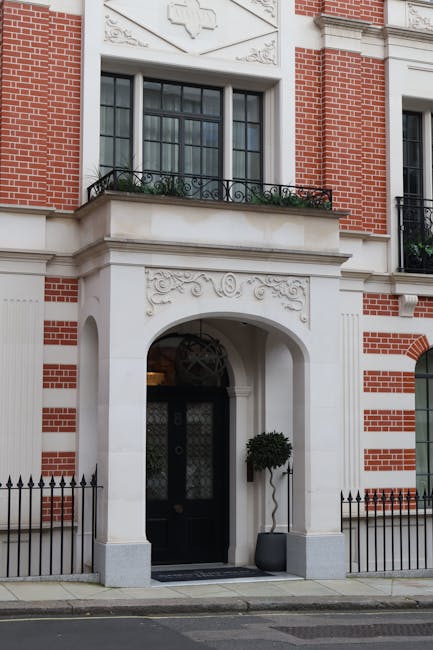 The image depicts the exterior of a historic red brick building in Mayfair, featuring white decorative stone accents and ornate carvings above the entrance. A black door with decorative glass panels is set within an arched white stone frame, flanked by a potted topiary plant. The building has large, multi-pane sash windows with black framing and small balconies with wrought iron railings. The pavement in front of the entrance is clean and well-maintained. Natural daylight illuminates the facade, highlighting the intricate architectural details. As part of their comprehensive domestic cleaning services, Cleaners Mayfair specializes in surface cleaning, deep cleaning, and sanitisation of historic and residential properties like this, ensuring the property's exterior remains pristine and well-maintained.