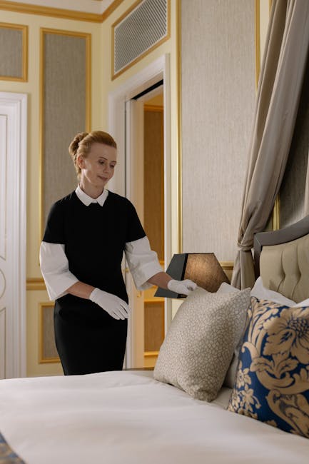 A professional housekeeper from Cleaners Mayfair, dressed in a black uniform with white gloves, is preparing a bedroom in a luxurious residence. She is standing beside a neatly made bed with white linens, and decorative pillows in beige and blue floral patterns. The room features elegant cream-colored walls with gold accents and paneling, with a large window draped with beige curtains. The housekeeper's hand rests on a small bedside table, possibly adjusting it or preparing for cleaning. The lighting is soft and warm, highlighting the clean, polished surfaces, including a cream upholstered headboard and a wooden frame around the window. The scene exemplifies meticulous surface cleaning, deep housekeeping, and routine sanitisation, reflecting the high standards of domestic cleaning services offered by Cleaners Mayfair in accordance with the Mayfair W1J Housekeeping Checklist for Berkeley Square, Mayfair.
