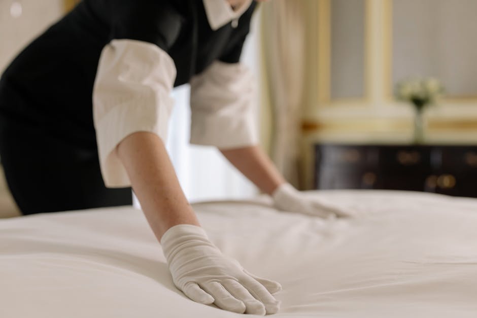 A professional cleaner from Cleaners Mayfair is shown in a well-lit bedroom, wearing white gloves and a black and white uniform, meticulously smoothing and straightening a white bed sheet on a neatly made bed. The bed features a wooden headboard, and in the background, there is a dark wooden dresser with a mirror and a decorative vase with white flowers placed on top. The room has a polished wooden floor, and the walls are painted in a light, neutral color, presenting a clean and hygienic environment suitable for deep cleaning and surface sanitisation inside a residential setting.