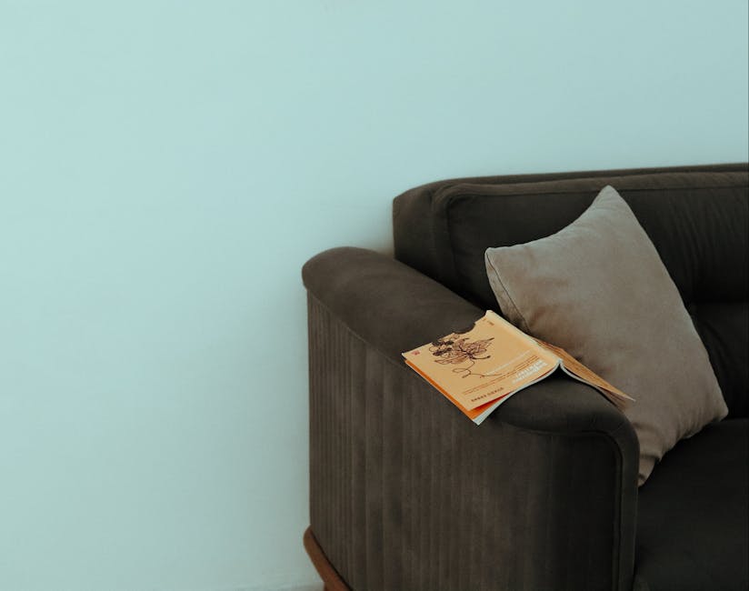 Close-up of a dark grey fabric sofa with a light grey cushion placed against its backrest, situated in a room with a pale mint green wall. A small stack of newspapers or magazines rests on the armrest of the sofa. The sofa appears clean and well-maintained, with no visible stains or dust, illustrating a setting suitable for surface cleaning or deep upholstery cleaning as part of a domestic cleaning service by Cleaners Mayfair. The scene is softly lit, highlighting the smooth texture of the fabric and the neat arrangement of objects, emphasizing hygiene and proper maintenance in a residential space.