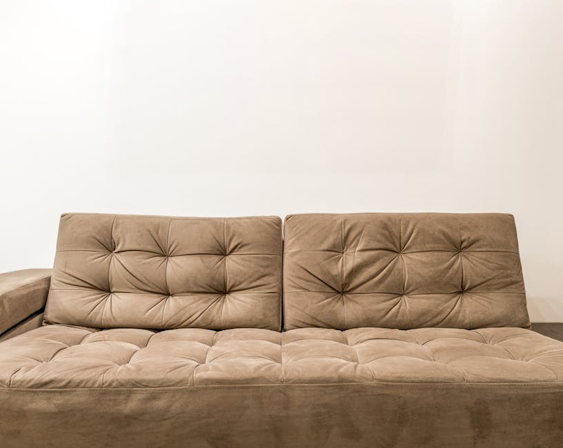 A beige fabric sofa with a tufted design on the cushions and backrest, positioned against a plain white wall in a tidy living room. The sofa has three seat sections and matching back cushions, with no visible stains or dirt, indicating recent deep cleaning and surface sanitisation by Cleaners Mayfair as part of their domestic cleaning services. The room is well-lit, showcasing the soft texture of the upholstery, and the floor is partially visible, appearing clean and uncluttered.
