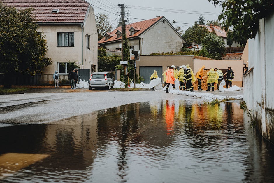 A group of workers wearing yellow and orange waterproof suits and helmets are engaged in flood cleanup on a residential street during daytime. They are positioned along the edge of a flooded area, with some handling large white sandbags or debris as part of surface cleaning and water removal efforts. The surrounding environment includes wet asphalt, puddles reflecting nearby houses with tiled roofs, and trees. In the background, residential buildings with white and grey facades, windows, and garage doors are visible, with utility poles and power lines overhead. The scene is illuminated by natural daylight, emphasizing the wet surfaces and the team's coordinated cleaning activities. This image illustrates a flood response effort by Cleaners Mayfair, demonstrating the importance of deep cleaning and sanitisation in flood-affected areas.