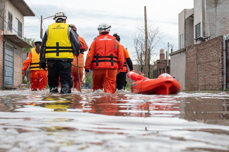 A group of rescue workers and emergency responders wearing orange and yellow high-visibility vests and helmets, walking through a flooded residential street with water covering the pavement. The surface of the water reflects the surrounding brick and concrete buildings, some with exposed brickwork and partially visible windows. An orange rescue boat is visible on the ground beside them, indicating flood response activity. The scene is set outdoors in a neighborhood with overcast skies, and the area appears to have experienced significant flooding, requiring urgent flood cleanup efforts, as managed by Cleaners Mayfair.