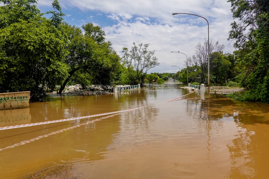 A group of workers wearing yellow and orange waterproof suits and helmets are engaged in flood cleanup on a residential street during daytime. They are positioned along the edge of a flooded area, with some handling large white sandbags or debris as part of surface cleaning and water removal efforts. The surrounding environment includes wet asphalt, puddles reflecting nearby houses with tiled roofs, and trees. In the background, residential buildings with white and grey facades, windows, and garage doors are visible, with utility poles and power lines overhead. The scene is illuminated by natural daylight, emphasizing the wet surfaces and the team's coordinated cleaning activities. This image illustrates a flood response effort by Cleaners Mayfair, demonstrating the importance of deep cleaning and sanitisation in flood-affected areas.